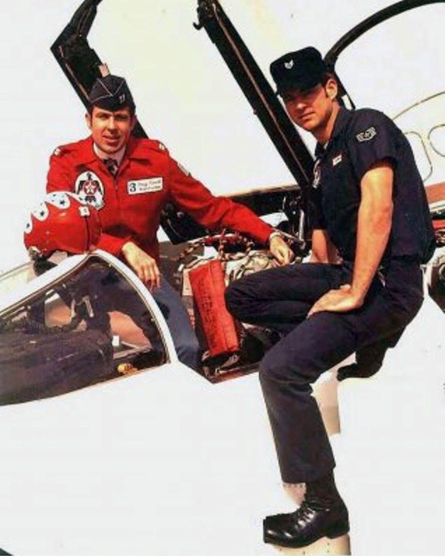 Captain Douglas Roach in a red Thunderbirds flight suit and Sergeant Jacobssen beside the cockpit of a Thunderbirds jet.